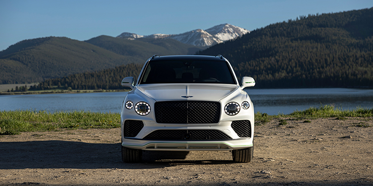 Bentley Bentayga Speed với ngoại thất Glacier White, nổi bật với lưới tản nhiệt màu đen bóng và cụm đèn pha pha lê đặc trưng, đỗ trước khung cảnh hồ nước và núi non yên bình – thể hiện sự tự tin và đẳng cấp trong từng chi tiết thiết kế.